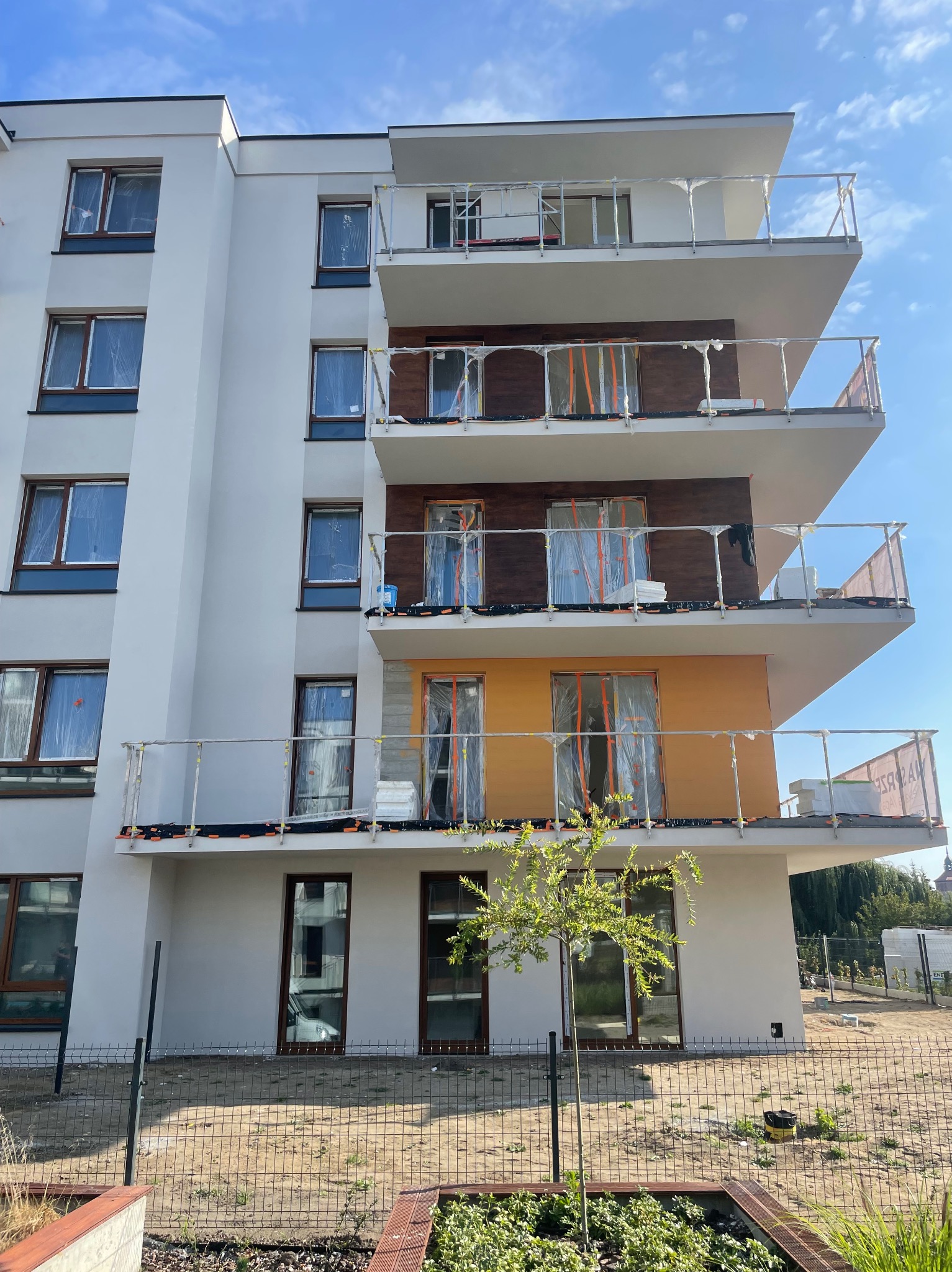 Wielopiętrowy budynek w trakcie wykańczania elewacji: białe ściany, fragmenty z drewnianą okładziną i pomarańczowym kolorem, zabezpieczone okna folią, balkony z szklanymi balustradami, widoczne...