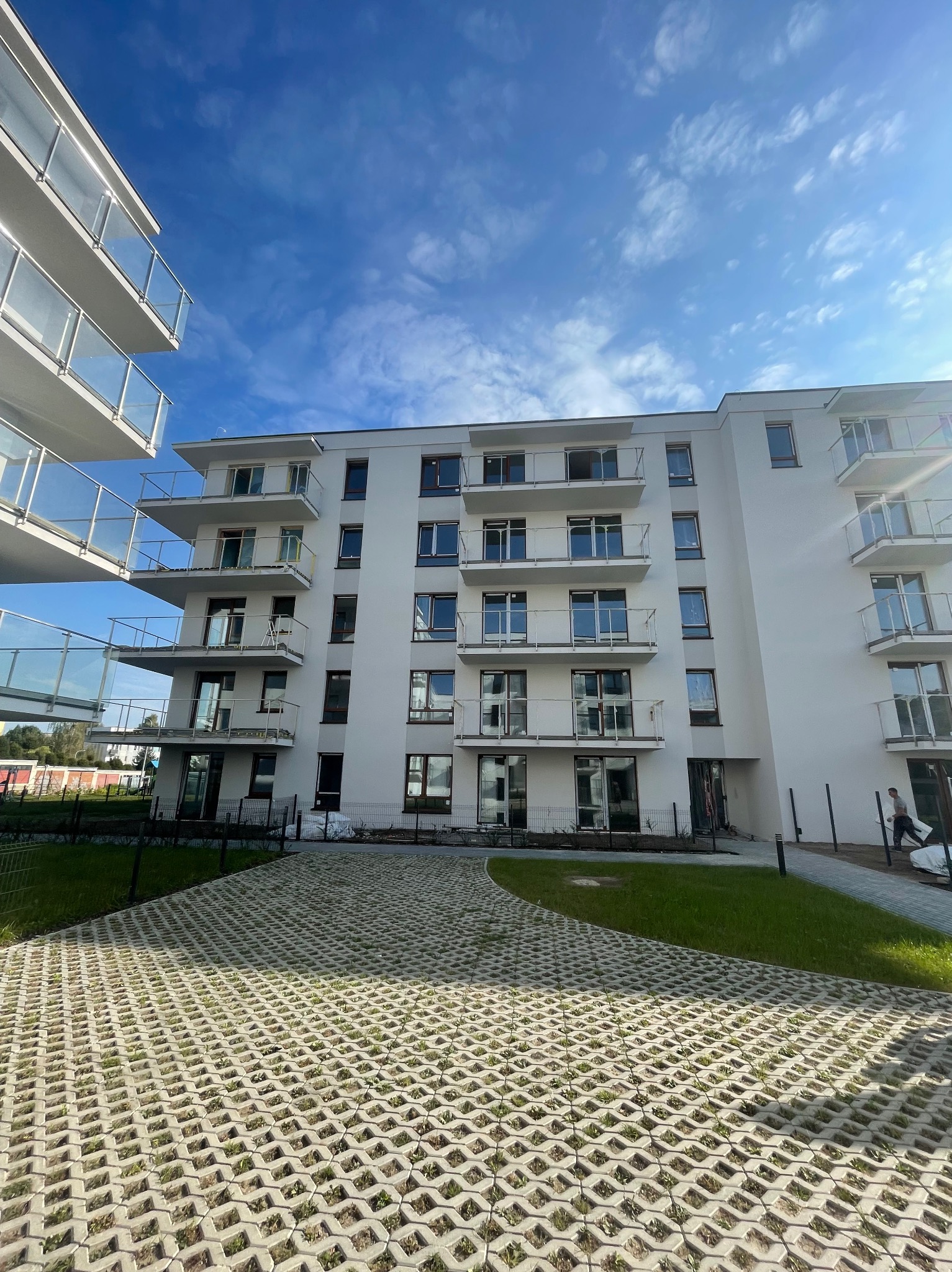 Nowoczesny, biały budynek wielorodzinny z balkonami o szklanych balustradach, widok z dołu na tle błękitnego nieba z chmurami, z przodu ażurowy parking z zielenią.