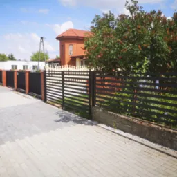 Nowoczesne, poziome ogrodzenie z czarnych metalowych profili, częściowo z podmurówką, przylegające do chodnika z szarej kostki brukowej, z budynkiem z czerwonej cegły w tle i zielenią drzew.