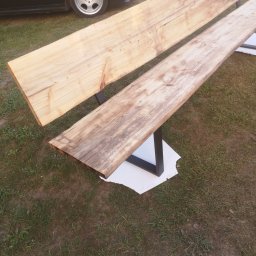 HUB-MAR Marek Piasecki - Dwie surowe deski z widocznym usłojeniem, ułożone równolegle na czarnych, metalowych nogach w kształcie litery U, na tle trawy i fragmentu czarnego samochodu.