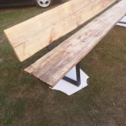 Dwie surowe deski z widocznym usłojeniem, ułożone równolegle na czarnych, metalowych nogach w kształcie litery U, na tle trawy i fragmentu czarnego samochodu.