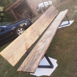 HUB-MAR Marek Piasecki - Dwa surowe blaty drewniane o nieregularnych krawędziach, oparate na metalowych, czarnych nogach w kształcie trapezu, ustawione na trawie obok czarnego samochodu i drewnianej wiaty.