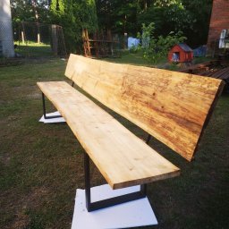 HUB-MAR Marek Piasecki - Drewniana ławka ogrodowa z oparciem wykonana z litego drewna o nieregularnych krawędziach, wsparta na czarnych, metalowych nogach w kształcie litery U, ustawiona na białych podkładkach na trawniku...