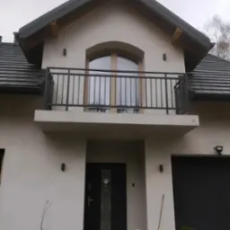 Elewacja domu z balkonem z czarną, metalową balustradą, widoczne czarne drzwi wejściowe i brama garażowa, dachówka w kolorze grafitowym.