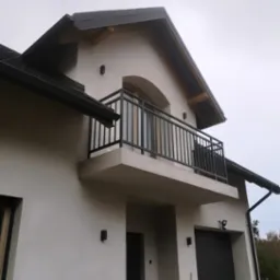 Nowoczesny balkon z czarną, metalową balustradą na tle białej fasady domu z szarym dachem.