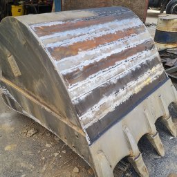 HUB-MAR Marek Piasecki - Ciężka, metalowa łyżka koparki po naprawie i spawaniu, widoczne ślady obróbki i nowe spoiny, leżąca na szutrowym podłożu w warsztacie.