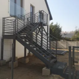 Zewnętrzne schody metalowe przy budynku z balustradą ze szkła. Schody prowadzą na balkon. Widoczne betonowe fundamenty i świeżo zagospodarowany teren wokół.