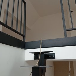 HUB-MAR Marek Piasecki - Nowoczesna, czarna balustrada metalowa z pionowymi szczeblami, zamocowana do białej ściany. Widoczne elementy mocujące i fragment konstrukcji nośnej.