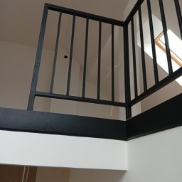 HUB-MAR Marek Piasecki - Minimalistyczna, czarna balustrada metalowa z pionowymi szczeblami, zamontowana na antresoli w jasnym wnętrzu. Widok z dołu, okno dachowe w tle.