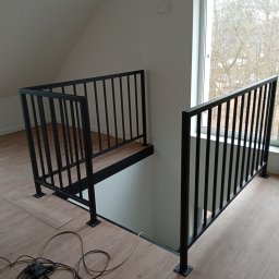 HUB-MAR Marek Piasecki - Nowoczesna, czarna balustrada metalowa na antresoli z drewnianym wykończeniem podłogi i dużym oknem w tle. Widoczne kable narzędzi na podłodze.