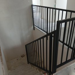 HUB-MAR Marek Piasecki - Nowoczesna, czarna balustrada metalowa na schodach w surowym stanie. Minimalistyczny design, pionowe pręty. Widok z góry, surowe wykończenie ścian i schodów.