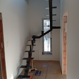 HUB-MAR Marek Piasecki - Nowoczesne, czarne schody metalowe wspornikowe w jasnym wnętrzu domu w Sędziejowicach. Widoczne elementy montażowe i zabezpieczenie podłogi kartonem.