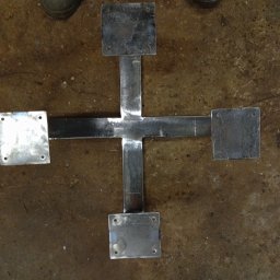 HUB-MAR - Metalowa konstrukcja w kształcie krzyża z przyspawanymi kwadratowymi płytkami, widoczne ślady spawania, szare buty robocze w tle, surowy wygląd.