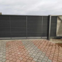 Nowoczesna, przesuwna brama wjazdowa z poziomymi panelami, pomalowana na grafitowo, zamontowana na betonowym fundamencie z kostką brukową w kształcie koniczyny.