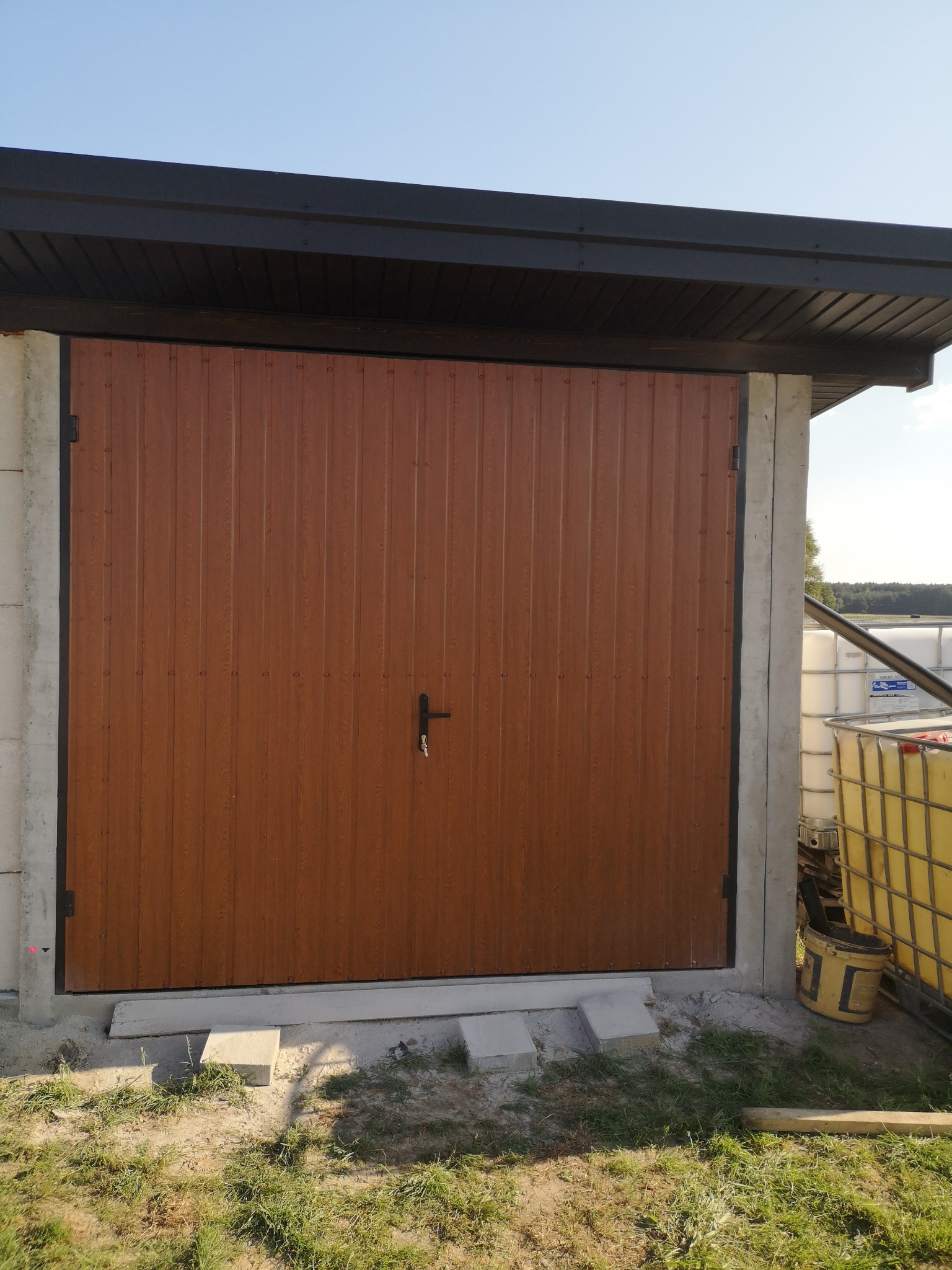 Brązowa brama garażowa z pionowymi panelami, zamontowana w surowym, betonowym otworze, widoczne betonowe płyty przed bramą i zbiorniki IBC w tle.