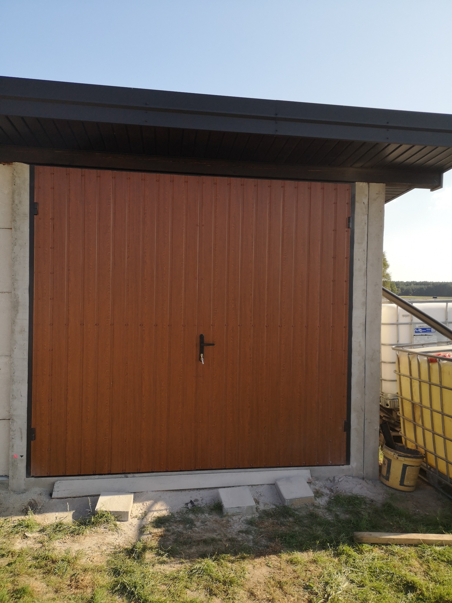 Zamknięta, brązowa brama garażowa z pionowymi panelami, zamontowana w surowej betonowej ramie, z widocznymi zbiornikami na wodę w tle.