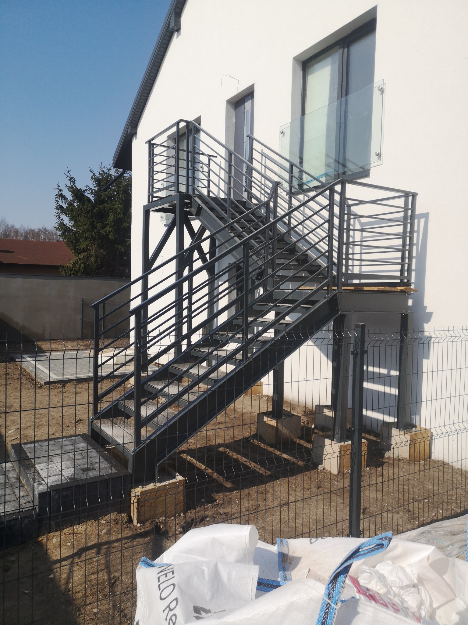 Zewnętrzne schody metalowe z balustradą, prowadzące na balkon z szklaną barierką, zamontowane przy białym budynku jednorodzinnym, widoczne przez ogrodzenie z siatki.