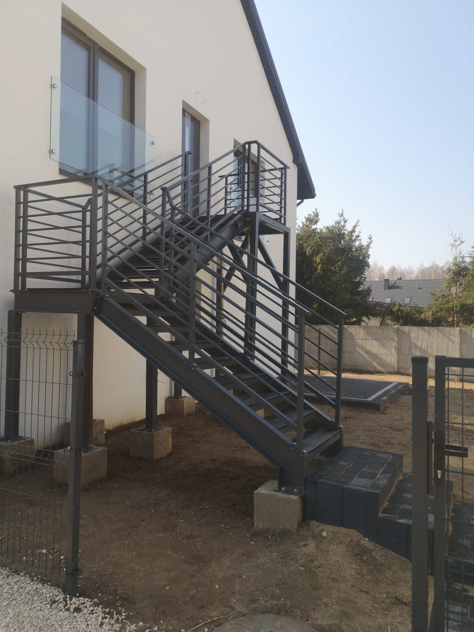 Zewnętrzne schody metalowe z balustradą, prowadzące na balkon z szklaną barierką, zamontowane przy elewacji budynku.