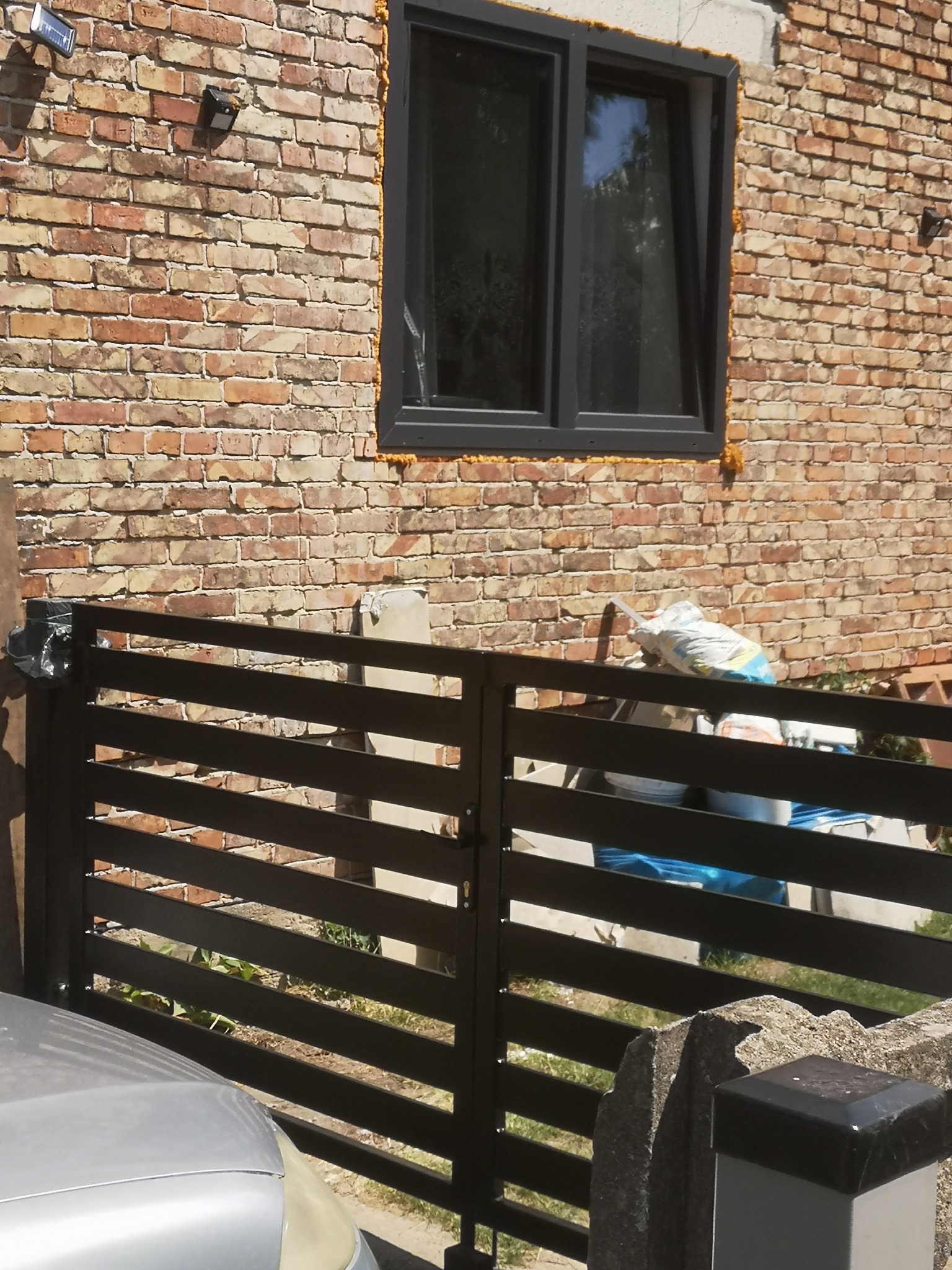 Nowoczesna, pozioma brama wjazdowa z metalowymi lamelami w kolorze antracytowym, na tle ceglanej ściany budynku z oknem z czarną ramą. Widoczne materiały budowlane i fragment srebrnego samochodu.