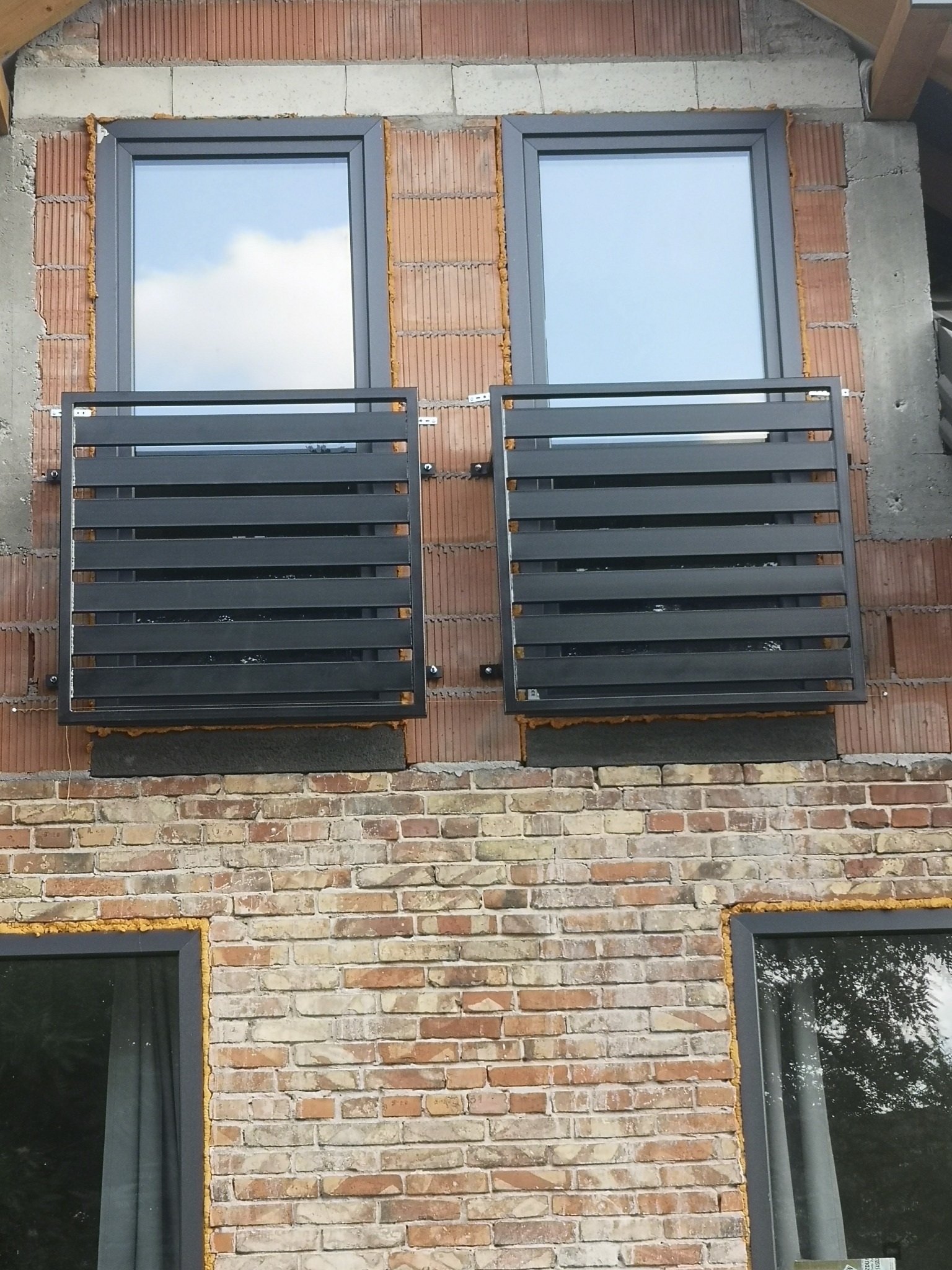 Zamontowane, czarne balustrady okienne z poziomych profili na ceglanej ścianie budynku w trakcie budowy, widoczne pianowanie okien.