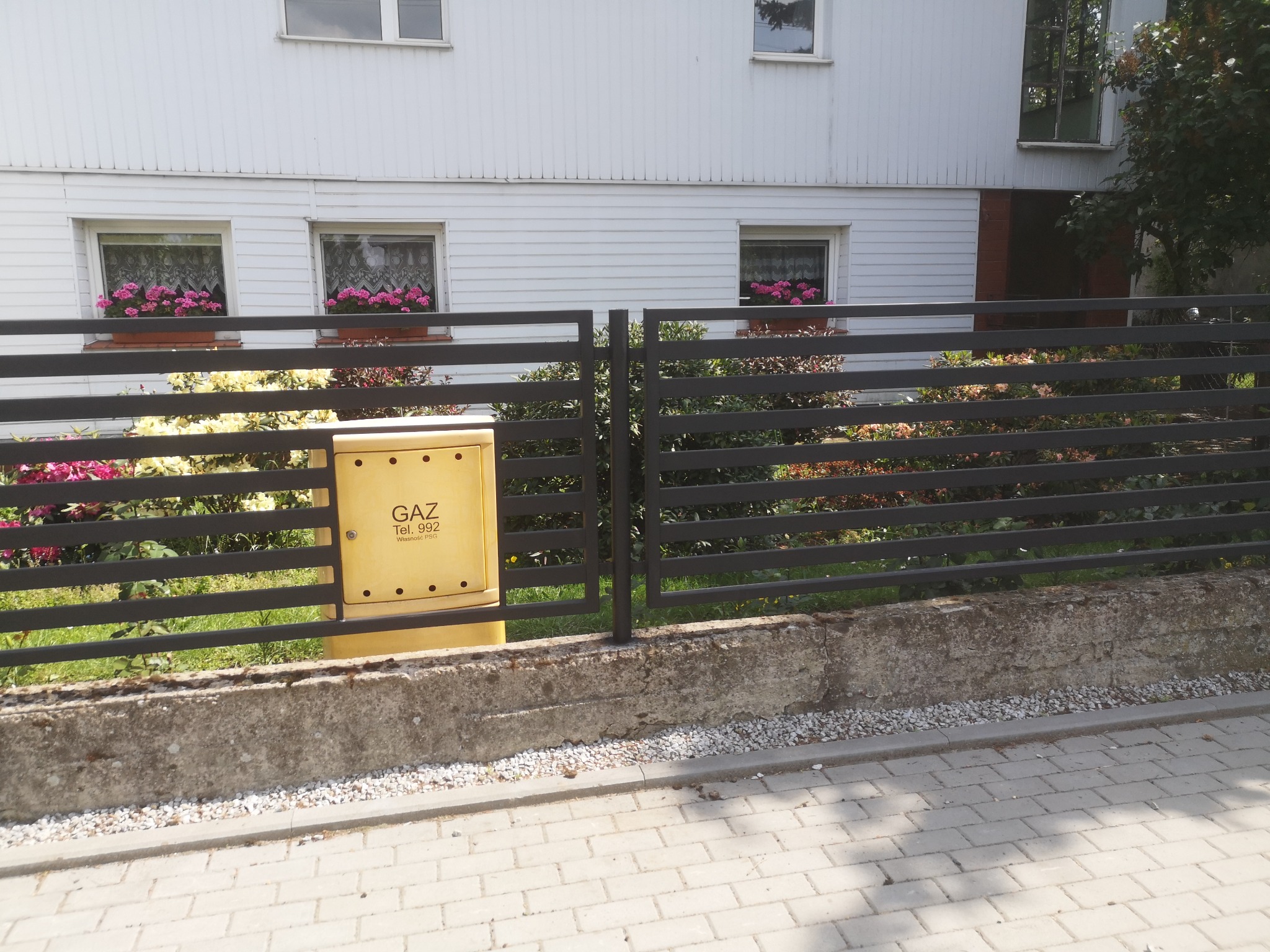 Nowoczesne ogrodzenie panelowe w kolorze antracytowym, z żółtą skrzynką gazową po lewej stronie, na tle białego domu z kwiatami w oknach.