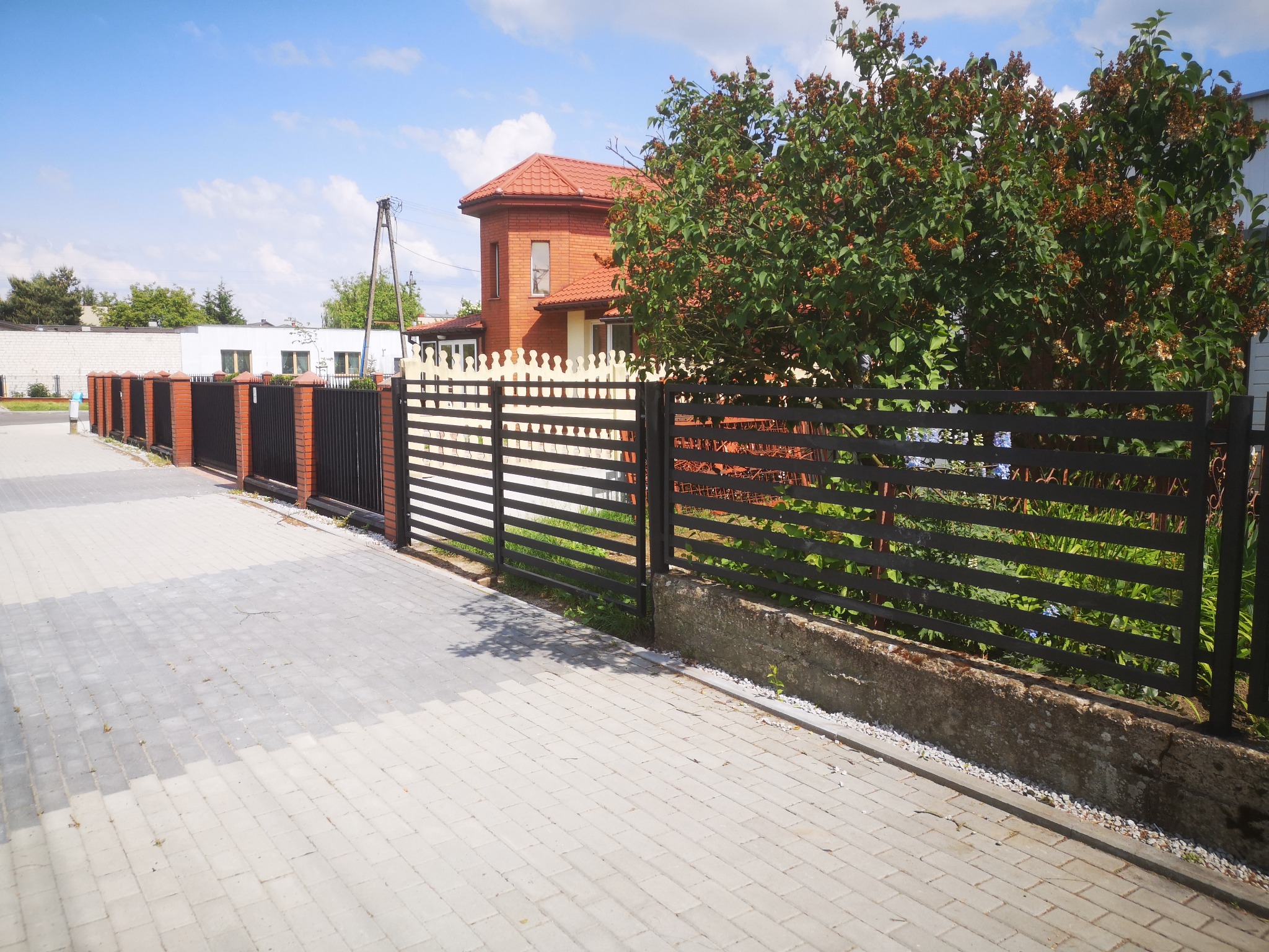 Nowoczesne, poziome ogrodzenie z czarnych metalowych profili, częściowo z podmurówką, przylegające do chodnika z szarej kostki brukowej, z budynkiem z czerwonej cegły w tle i zielenią drzew.