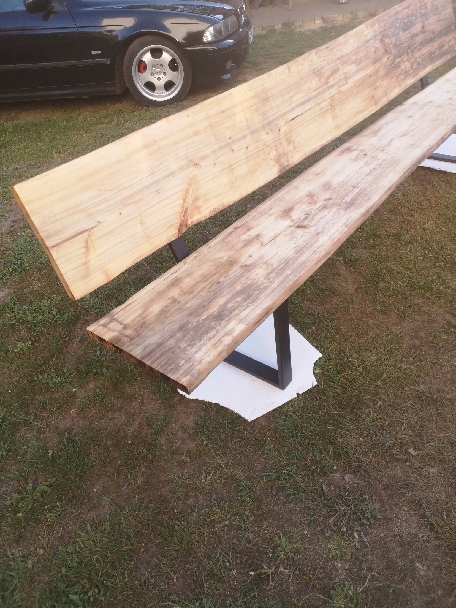 Dwie surowe deski z widocznym usłojeniem, ułożone równolegle na czarnych, metalowych nogach w kształcie litery U, na tle trawy i fragmentu czarnego samochodu.