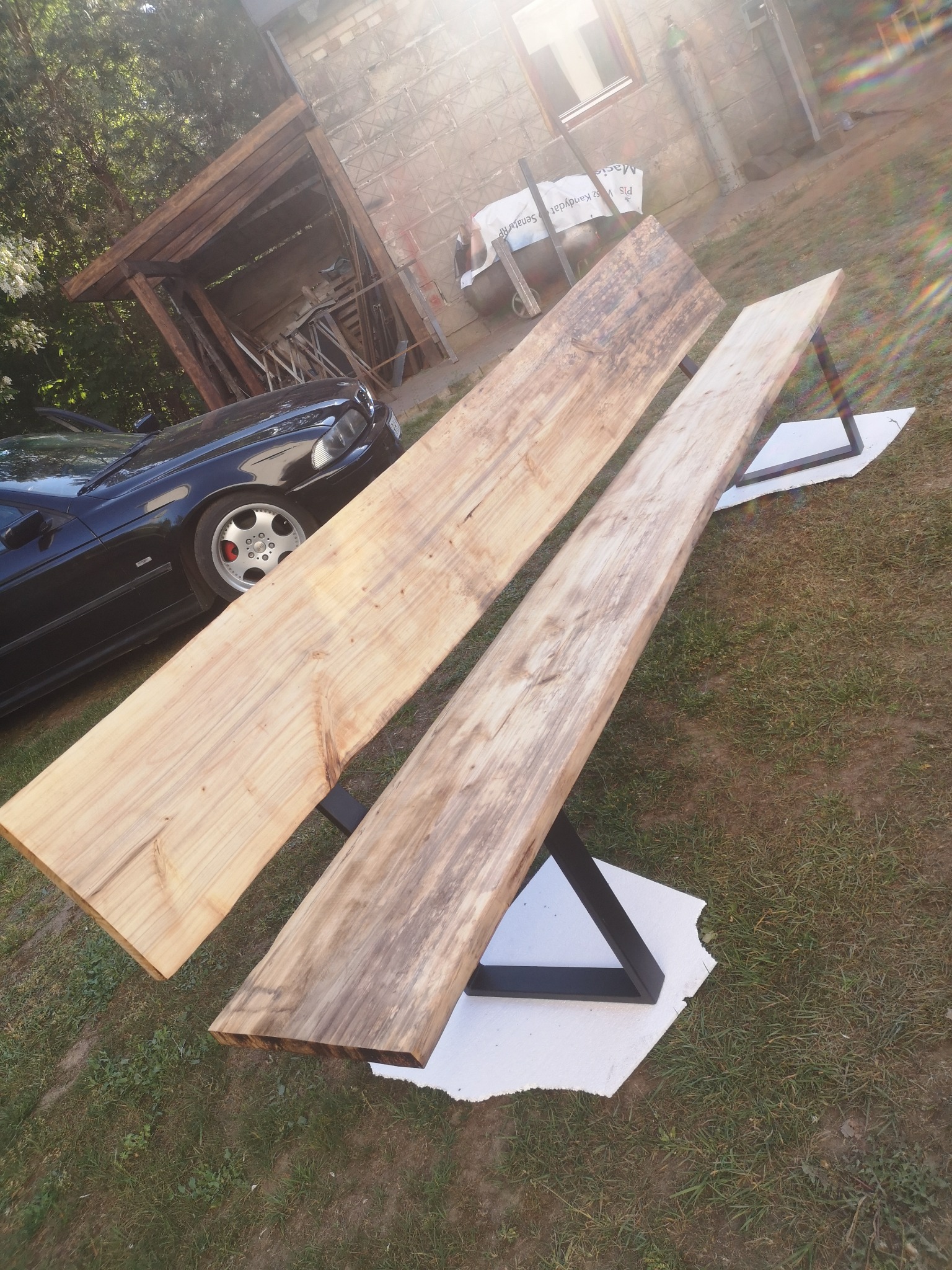 Dwa surowe blaty drewniane o nieregularnych krawędziach, oparate na metalowych, czarnych nogach w kształcie trapezu, ustawione na trawie obok czarnego samochodu i drewnianej wiaty.