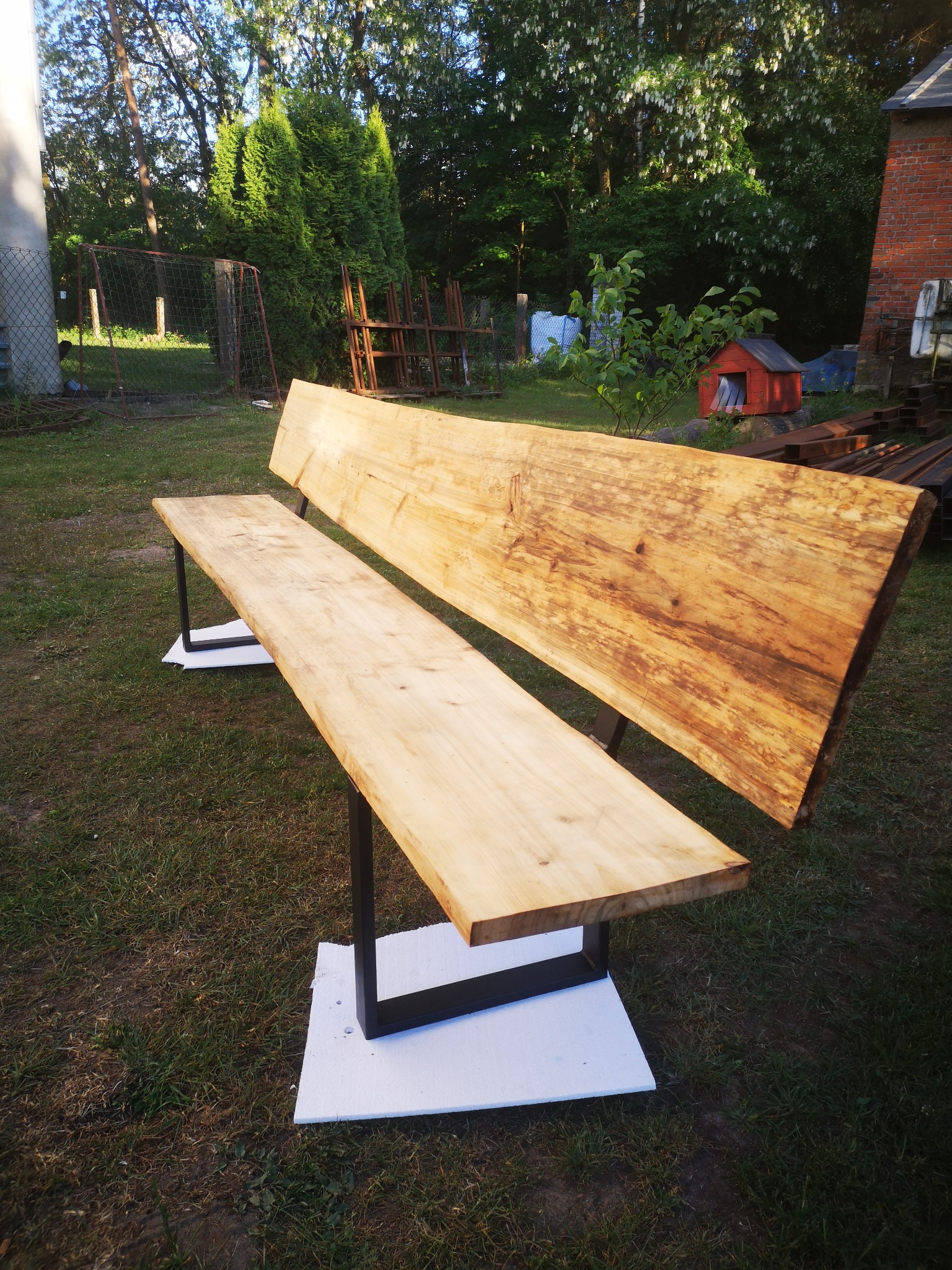 Drewniana ławka ogrodowa z oparciem wykonana z litego drewna o nieregularnych krawędziach, wsparta na czarnych, metalowych nogach w kształcie litery U, ustawiona na białych podkładkach na trawniku...