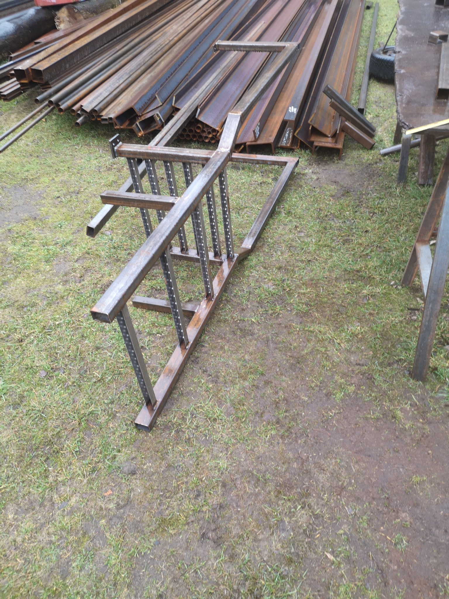 Element metalowej konstrukcji schodów w trakcie spawania, leżący na trawie obok stosu profili stalowych.