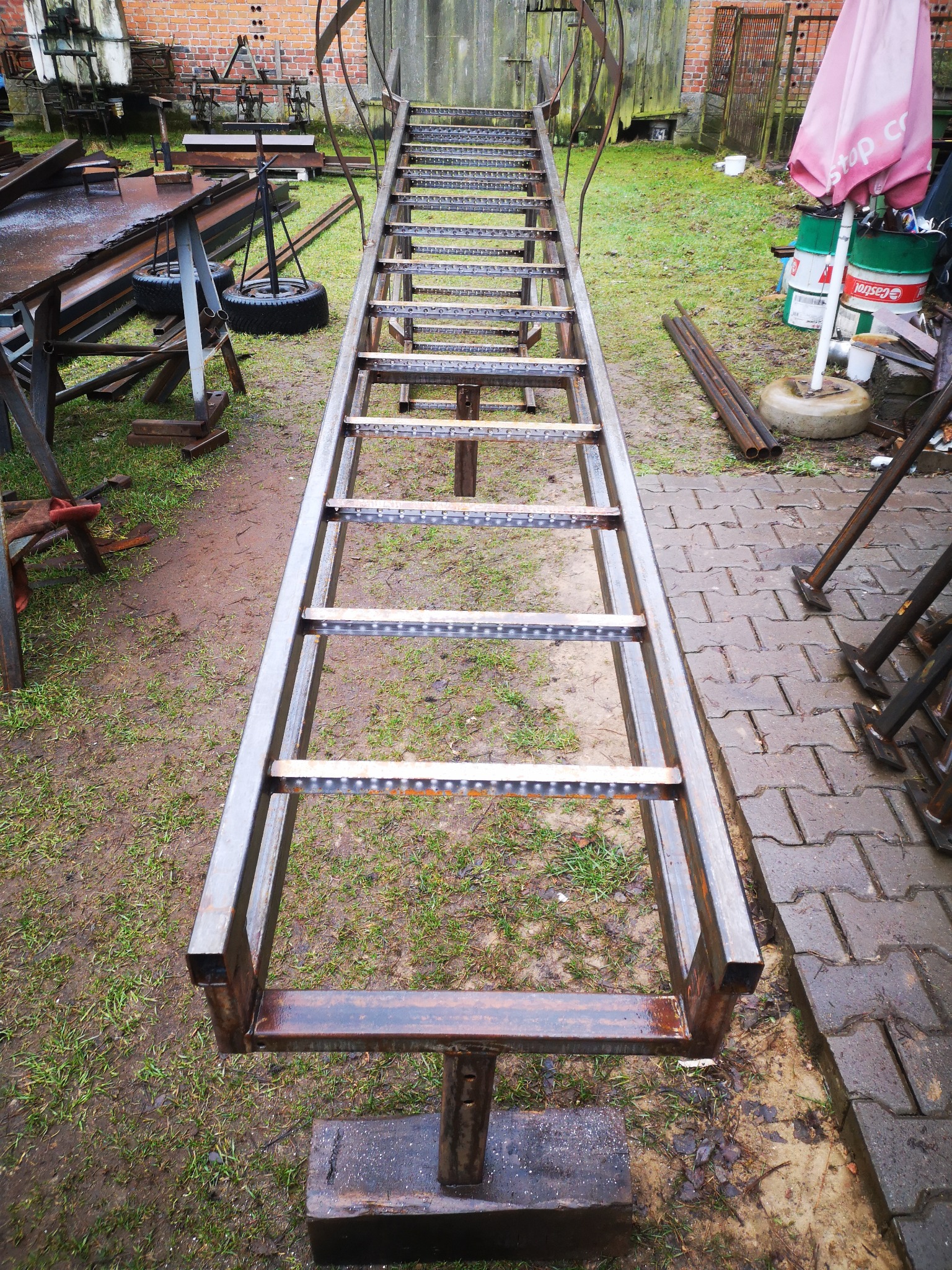 Metalowa konstrukcja schodów w trakcie budowy, widoczne elementy spawane, surowe wykończenie, tło z ceglanym budynkiem i zielonym trawnikiem.