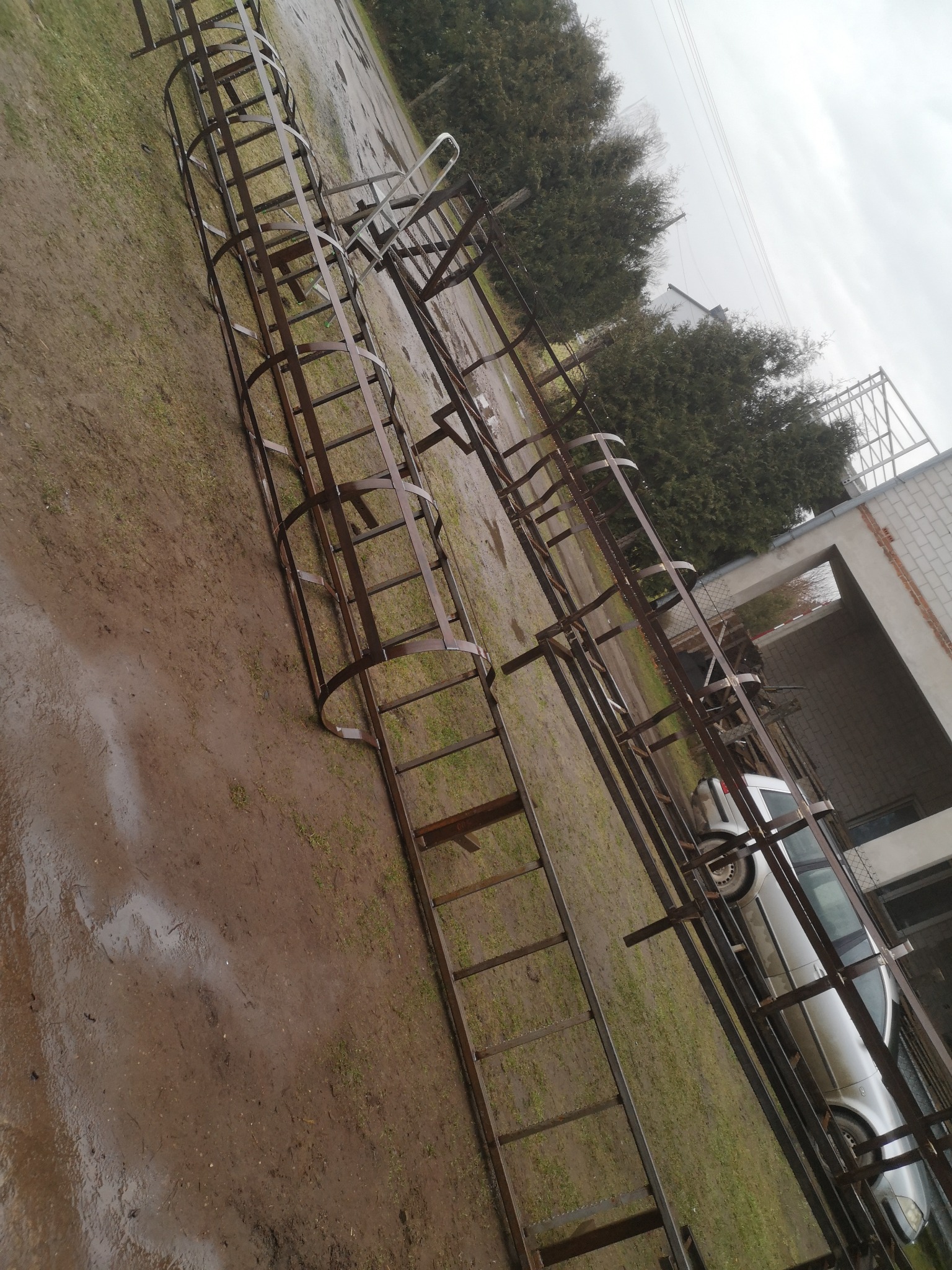 Surowa konstrukcja stalowa schodów metalowych z elementami okrągłymi, leżąca na mokrej trawie w pochmurny dzień, widoczny samochód w tle.