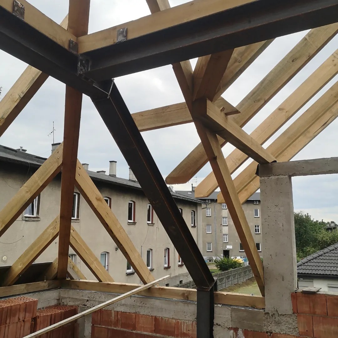 Wiązary dachowe z belek drewnianych i stalowych, widok z wnętrza budowy na tle budynków mieszkalnych.