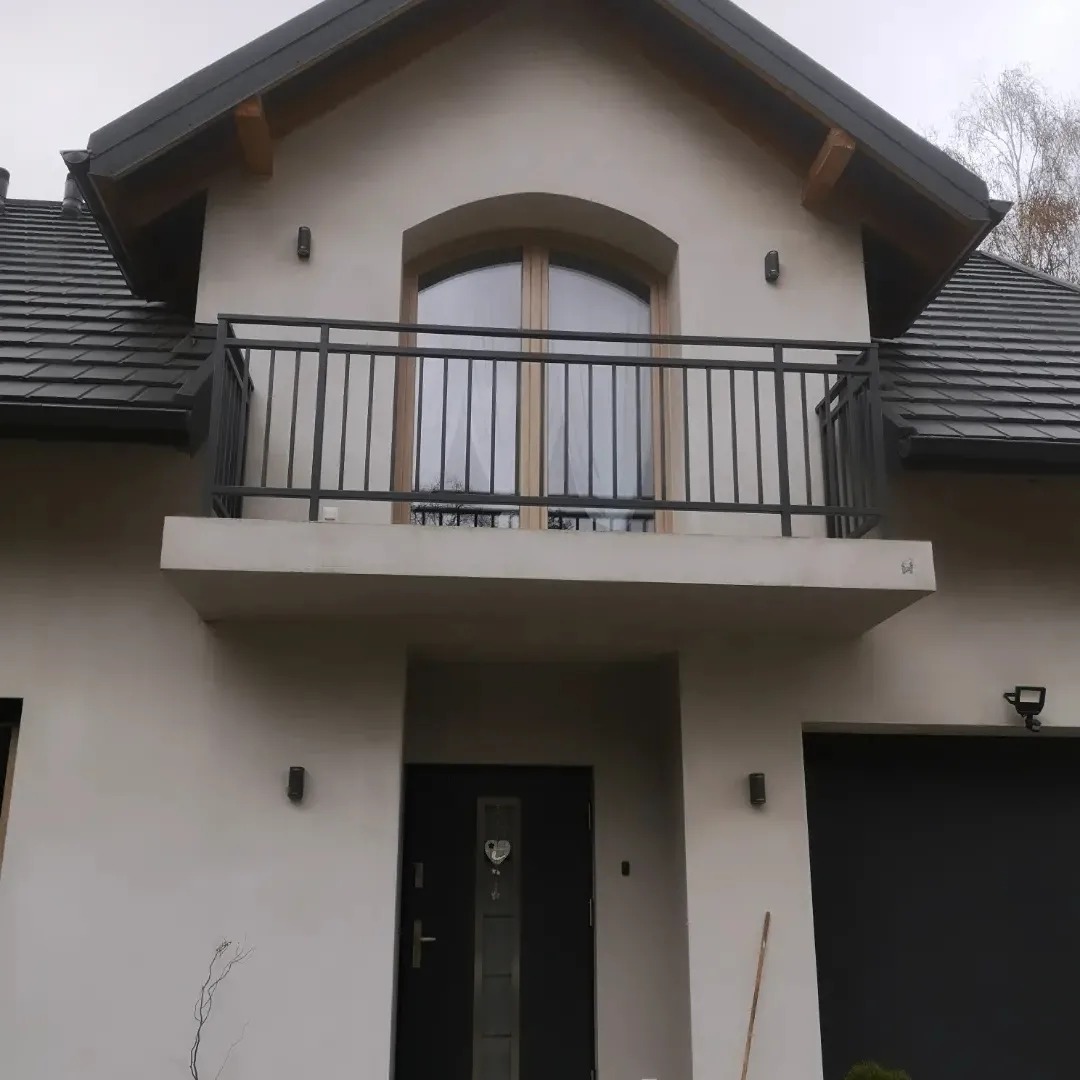Elewacja domu z balkonem z czarną, metalową balustradą, widoczne czarne drzwi wejściowe i brama garażowa, dachówka w kolorze grafitowym.