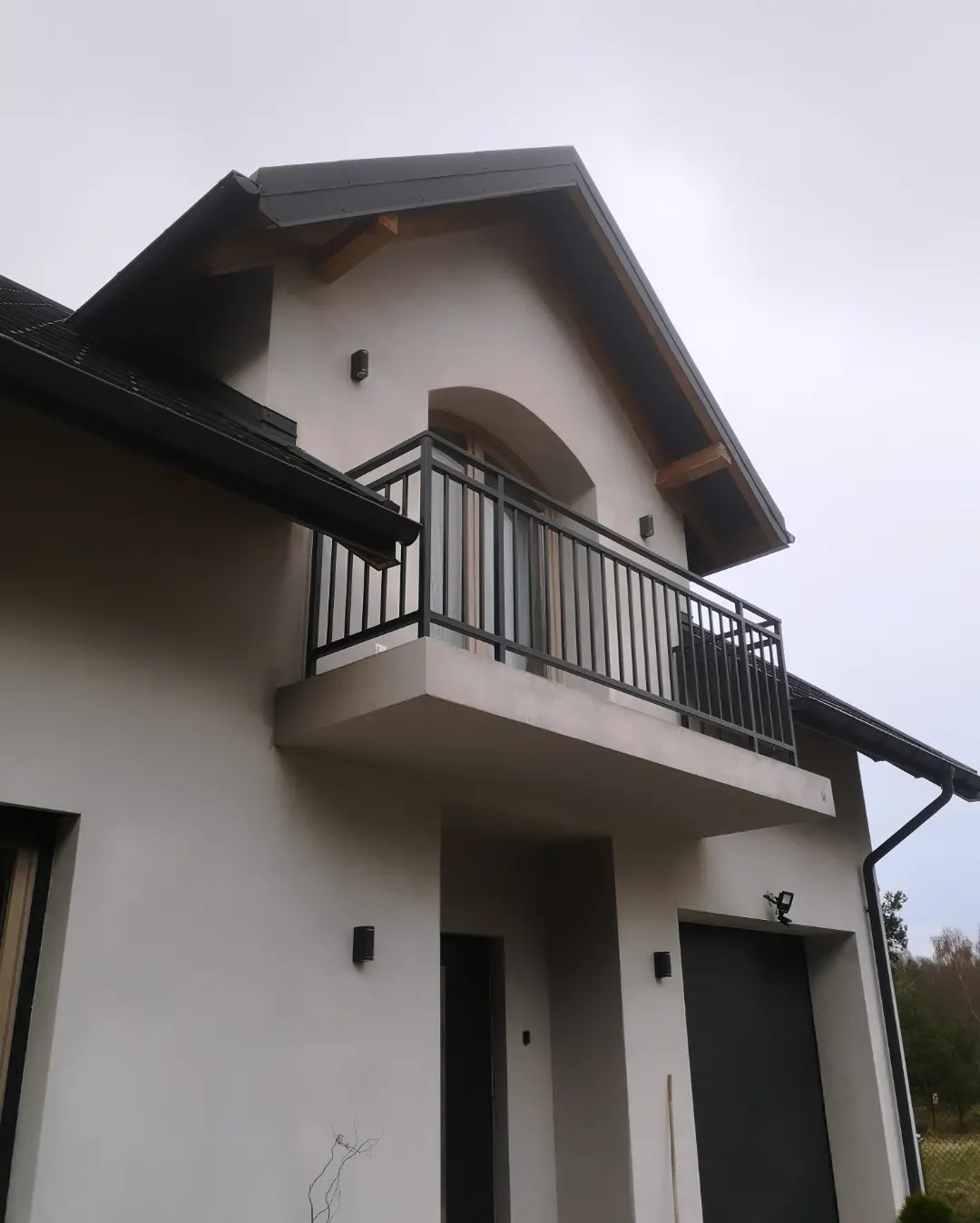 Nowoczesny balkon z czarną, metalową balustradą na tle białej fasady domu z szarym dachem.