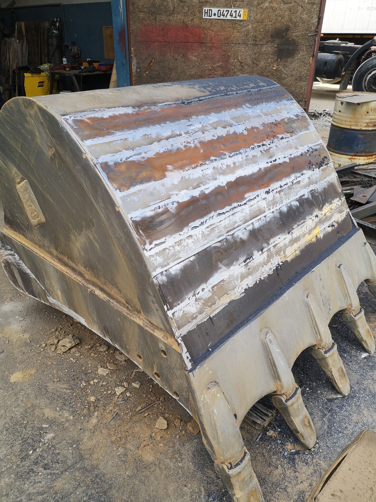 Ciężka, metalowa łyżka koparki po naprawie i spawaniu, widoczne ślady obróbki i nowe spoiny, leżąca na szutrowym podłożu w warsztacie.