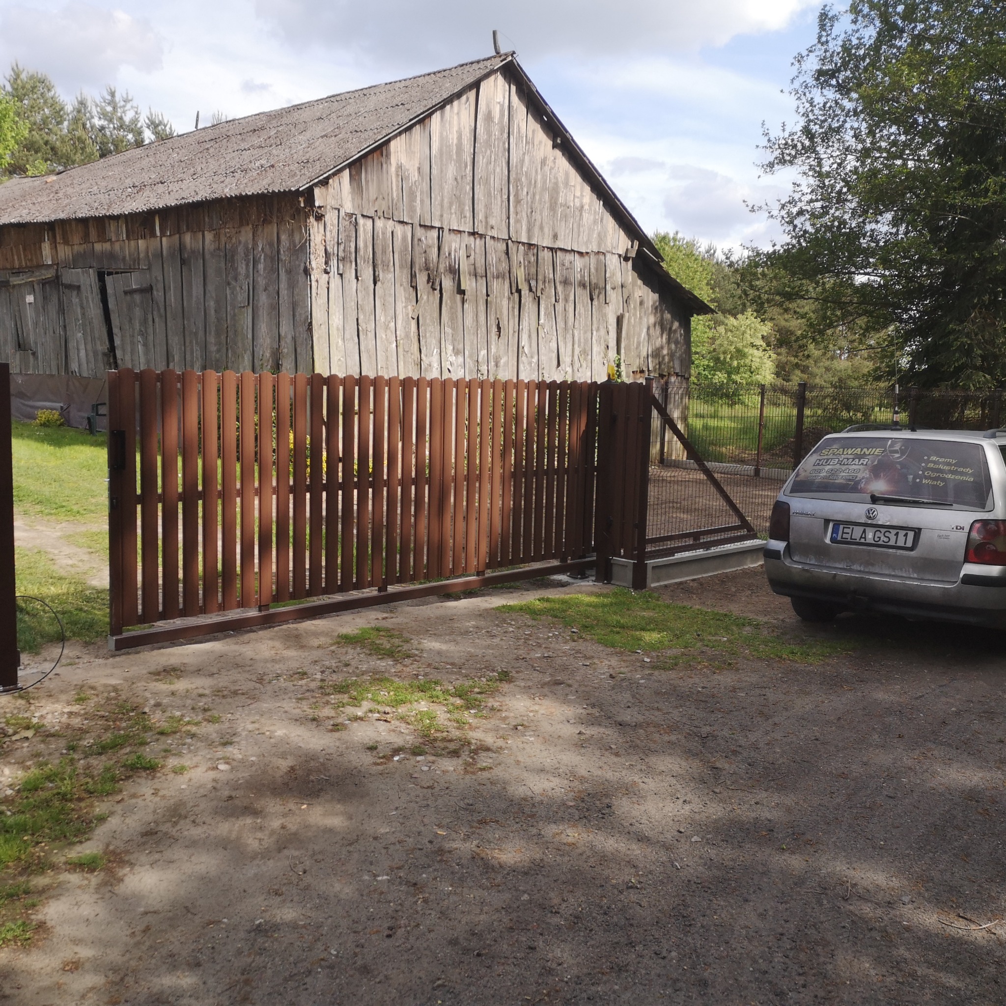 Brązowa brama wjazdowa z pionowych sztachet na tle starej, drewnianej stodoły i zaparkowanego auta z reklamą firmy. Widok z zewnątrz posesji.