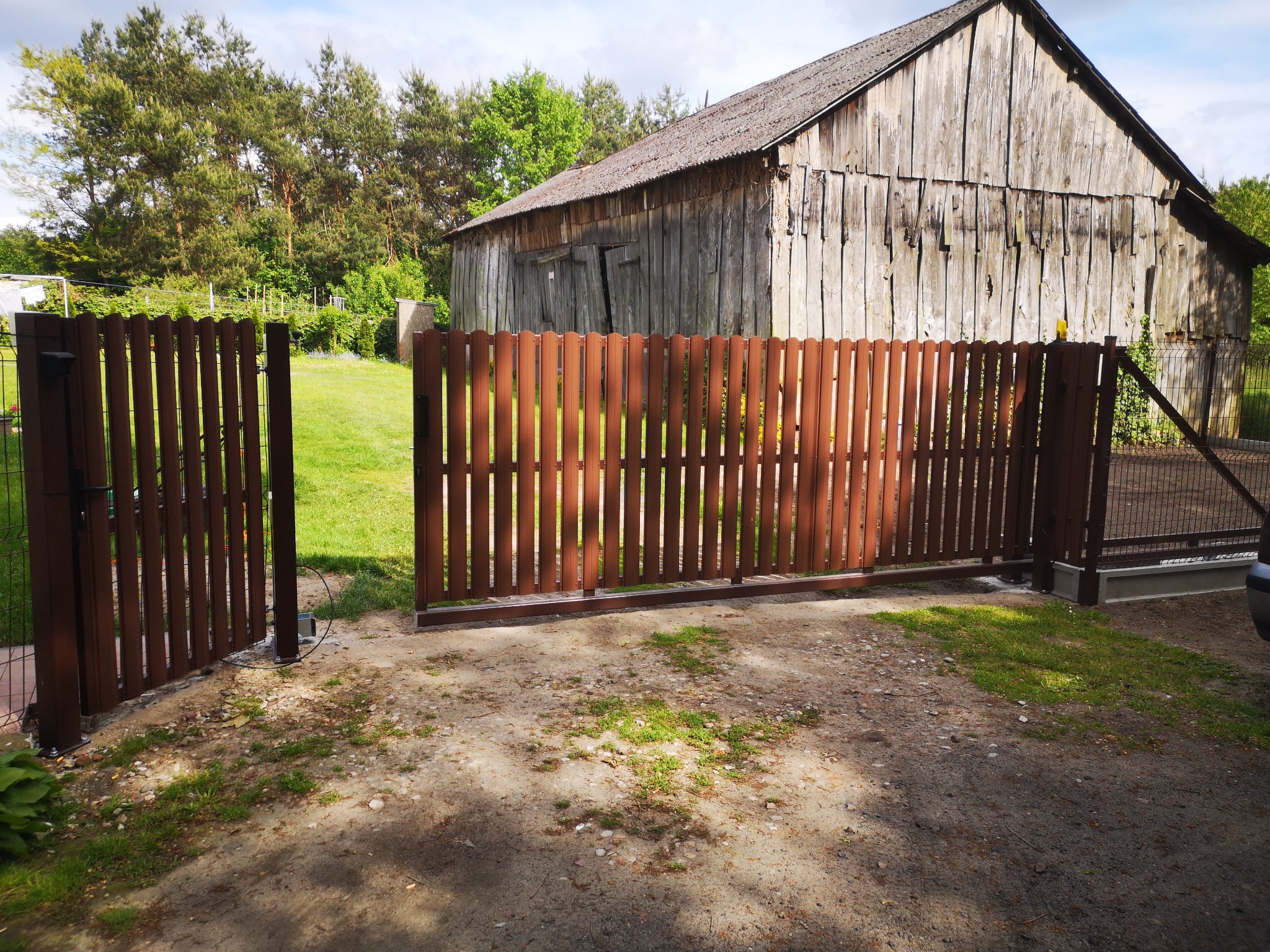 Otwarta, brązowa brama wjazdowa z pionowych sztachet na tle starej, drewnianej stodoły i zieleni. Widoczny mechanizm otwierania. Brama z metalowymi słupkami.