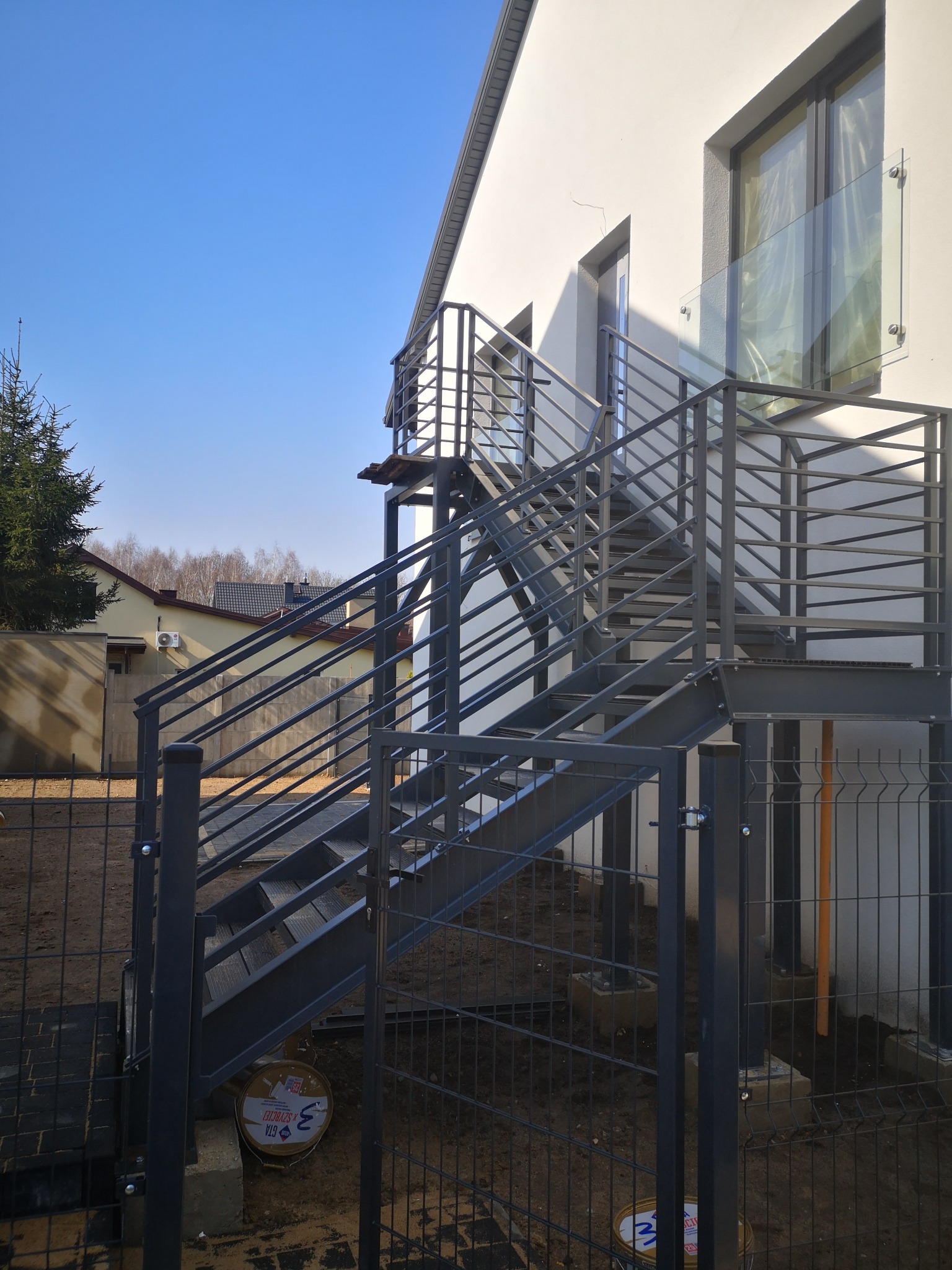 Zewnętrzne schody metalowe z balustradą, prowadzące do wejścia na piętro budynku. Widoczne ogrodzenie panelowe i fragment elewacji z oknem zabezpieczonym szkłem.