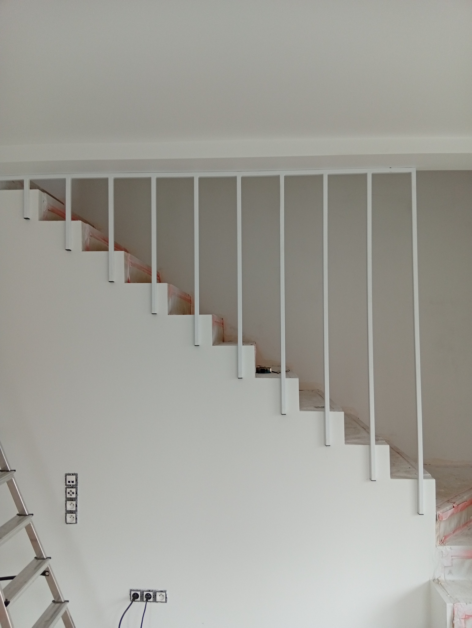Białe schody z metalową balustradą w minimalistycznym stylu, widoczne gniazdka elektryczne i drabina, sugerujące prace wykończeniowe.