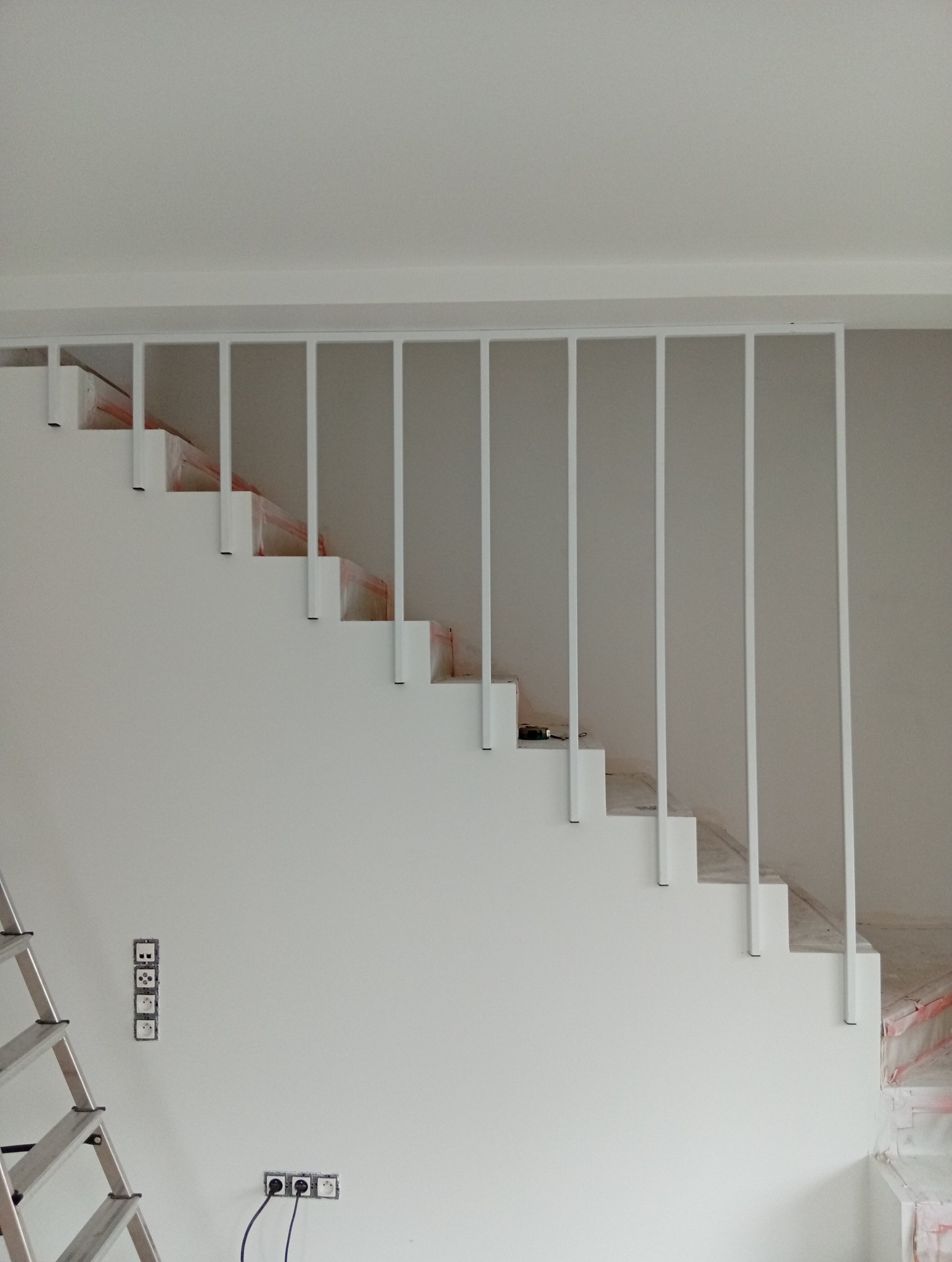 Białe schody z metalową balustradą w minimalistycznym stylu, widoczne gniazdka elektryczne i drabina, wnętrze w trakcie wykończenia.