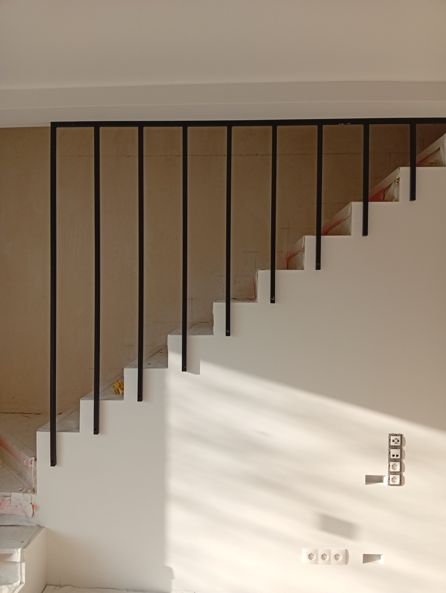 Nowoczesne schody z czarną, metalową balustradą w minimalistycznym wnętrzu. Widoczne gniazdka elektryczne na ścianie, częściowo w stanie surowym.