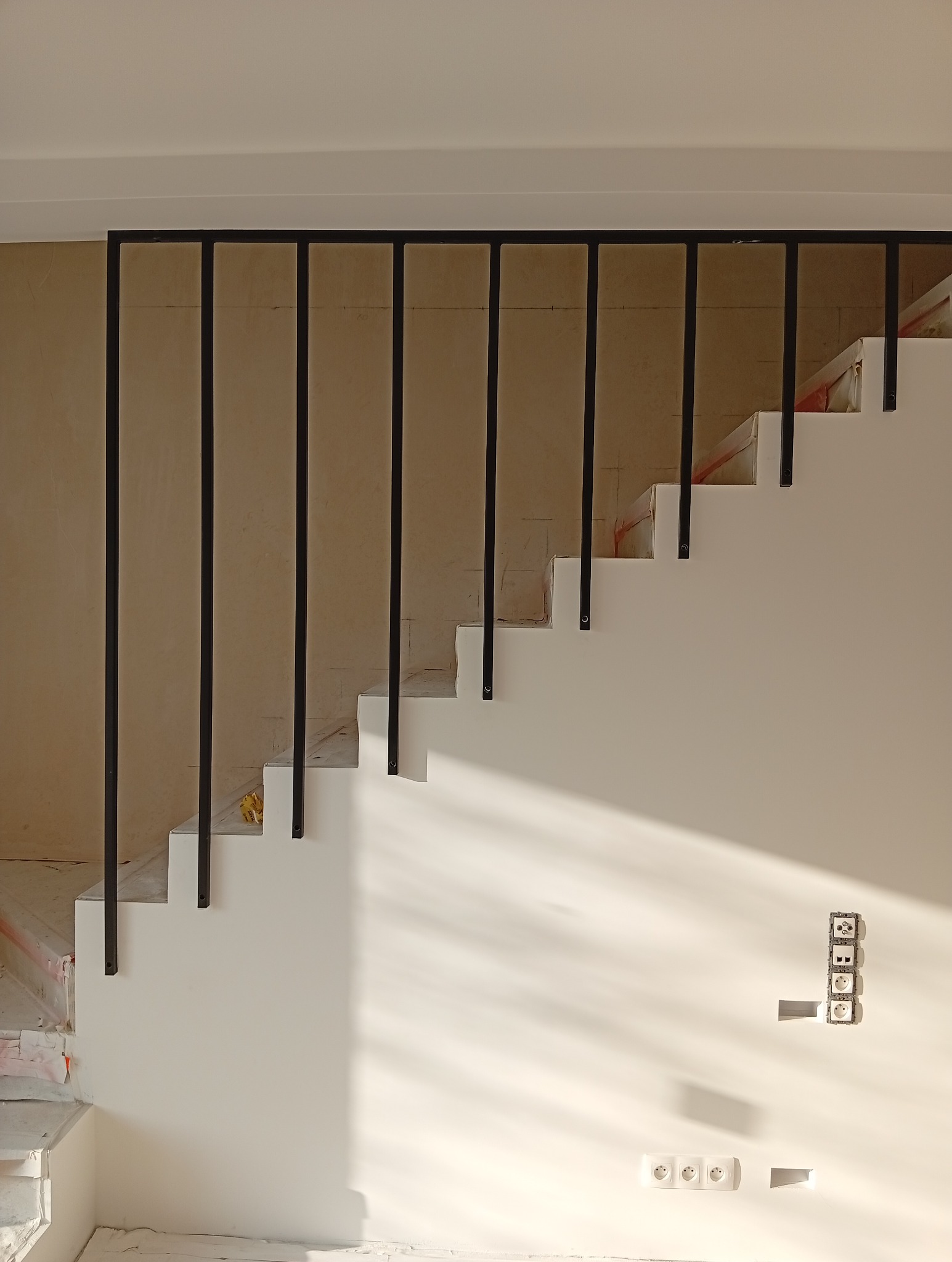 Nowoczesne schody z czarną, metalową balustradą w jasnym wnętrzu. Widoczne gniazdka elektryczne i cień. Minimalistyczny design, surowe wykończenie.