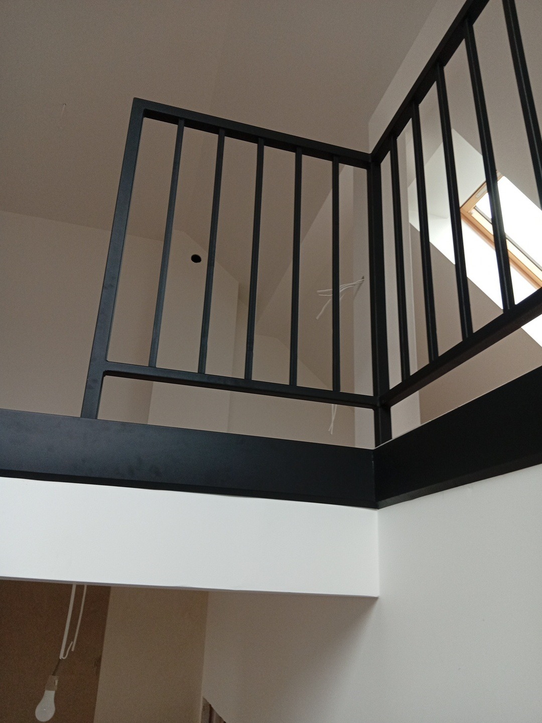 Minimalistyczna, czarna balustrada metalowa z pionowymi szczeblami, zamontowana na antresoli w jasnym wnętrzu. Widok z dołu, okno dachowe w tle.