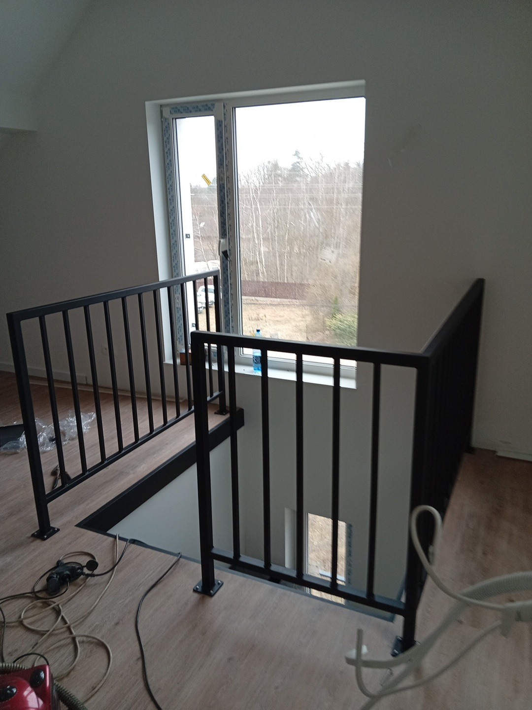 Nowoczesne, czarne schody metalowe z balustradą w jasnym wnętrzu. Widok z góry na dół, okno w tle. Na podłodze widoczne narzędzia i kable.