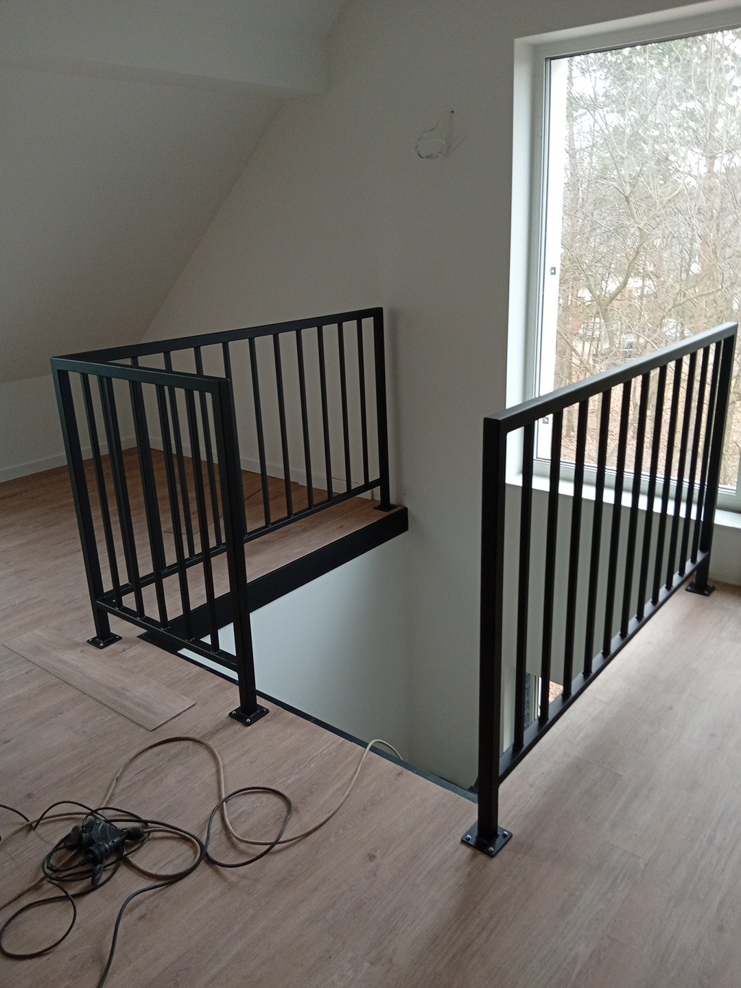 Nowoczesna, czarna balustrada metalowa na antresoli z drewnianym wykończeniem podłogi i dużym oknem w tle. Widoczne kable narzędzi na podłodze.