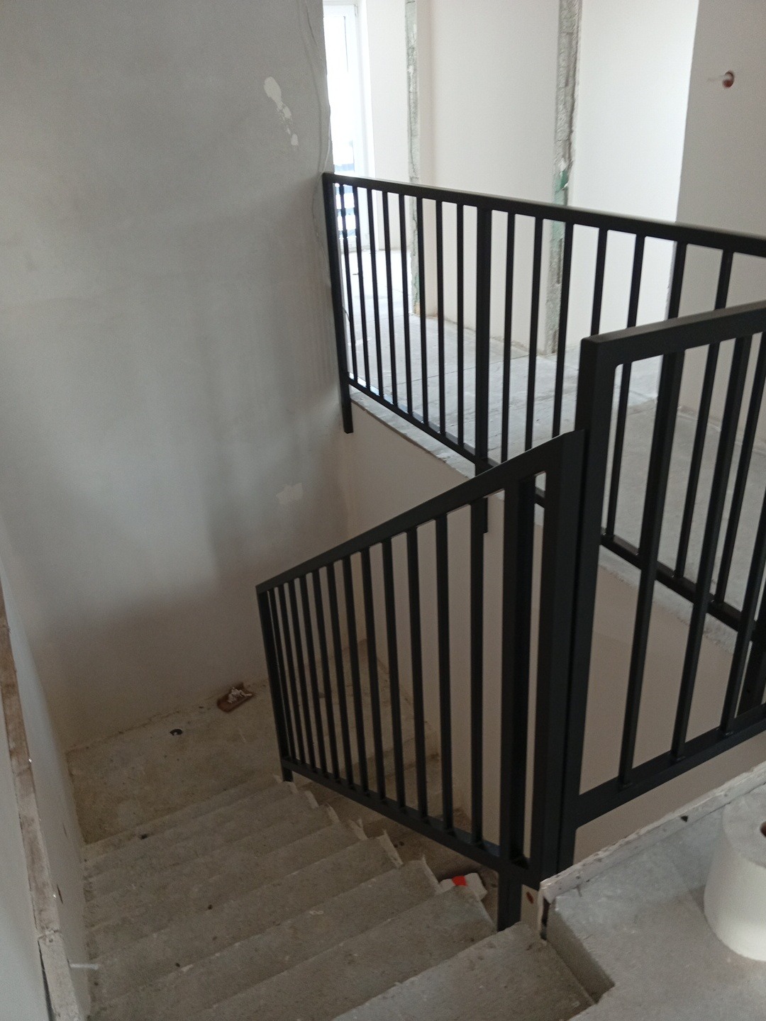 Nowoczesna, czarna balustrada metalowa na schodach w surowym stanie. Minimalistyczny design, pionowe pręty. Widok z góry, surowe wykończenie ścian i schodów.