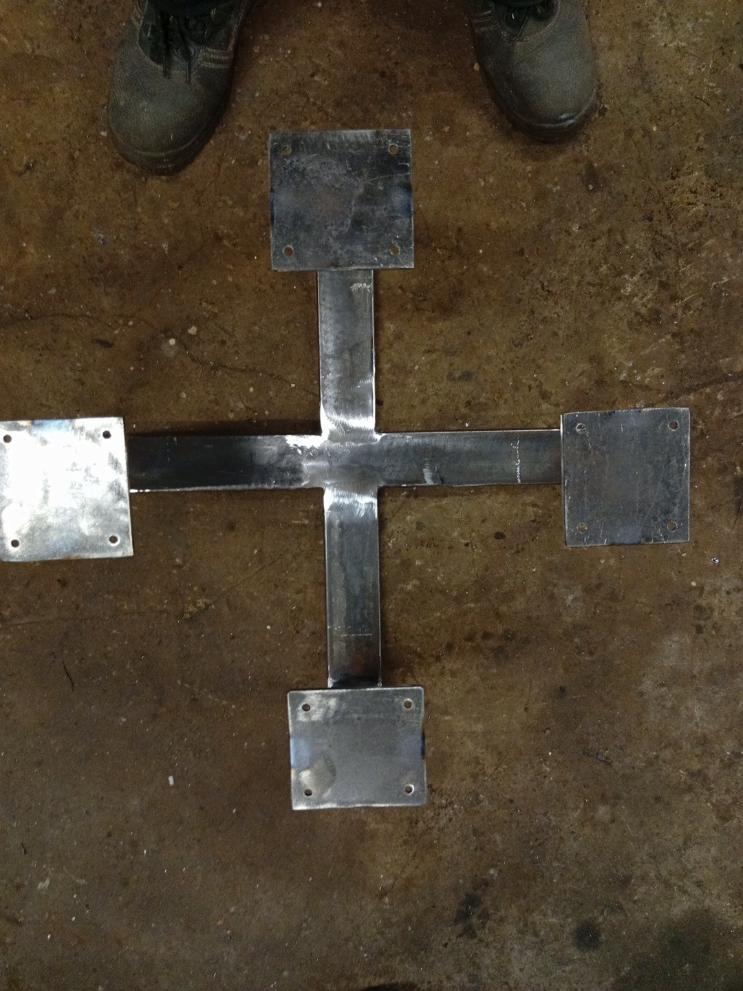 Metalowa konstrukcja w kształcie krzyża z przyspawanymi kwadratowymi płytkami, widoczne ślady spawania, szare buty robocze w tle, surowy wygląd.