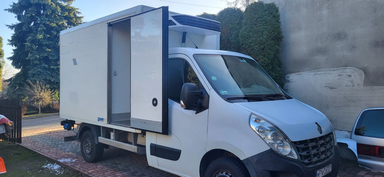 Biały samochód dostawczy typu chłodnia z otwartymi tylnymi drzwiami, zaparkowany na brukowanym podjeździe przed domem z zielonymi krzewami w tle.