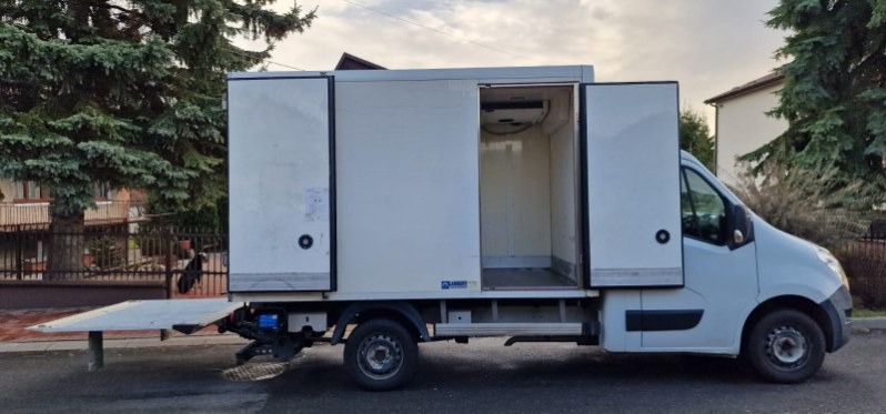 Biała furgonetka chłodnia z otwartymi drzwiami skrzydłowymi, widoczny mechanizm chłodniczy wewnątrz, platforma załadunkowa opuszczona, zaparkowana na ulicy.